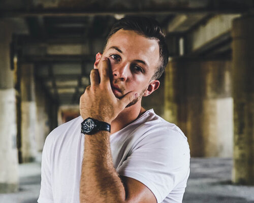 A man in a white t-shirt stands in an industrial setting, lightly touching his face with one hand. He wears a black watch and is positioned in front of concrete pillars, creating a moody and urban atmosphere.