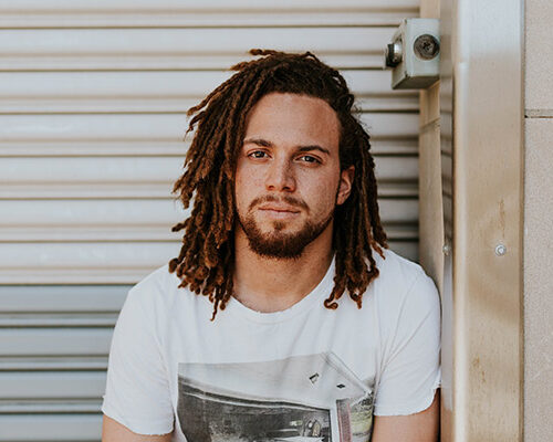 A man with long curly hair wearing a white t-shirt, sitting casually against a wall with a metal roll-up door in the background.