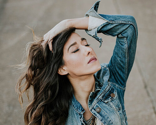 A woman with long, wavy hair wearing a denim jacket, posing outdoors with her hand on her head. She has her eyes closed and appears to be in a thoughtful pose against a neutral background.