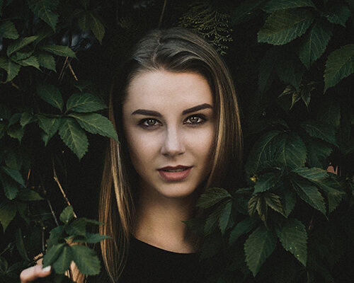 A woman with long brown hair standing among green foliage, looking directly at the camera. Her expression is neutral, and she is partially framed by leaves.
