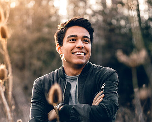 A young man in a dark jacket standing outdoors among tall plants, smiling while crossing his arms. The background shows a natural setting with soft sunlight filtering through trees.