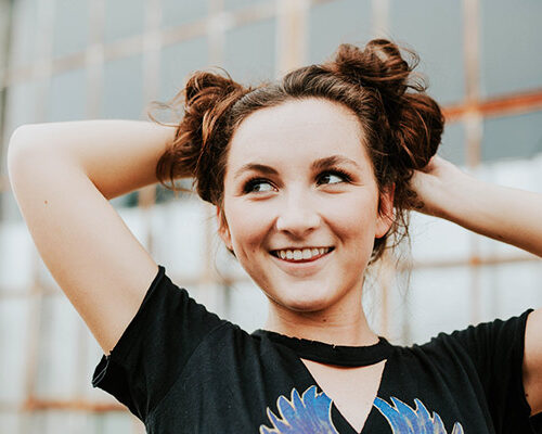 A young woman with styled hair in two buns is playfully arranging her hair while smiling. She wears a black t-shirt with a colorful design, and a blurred industrial background is visible behind her.