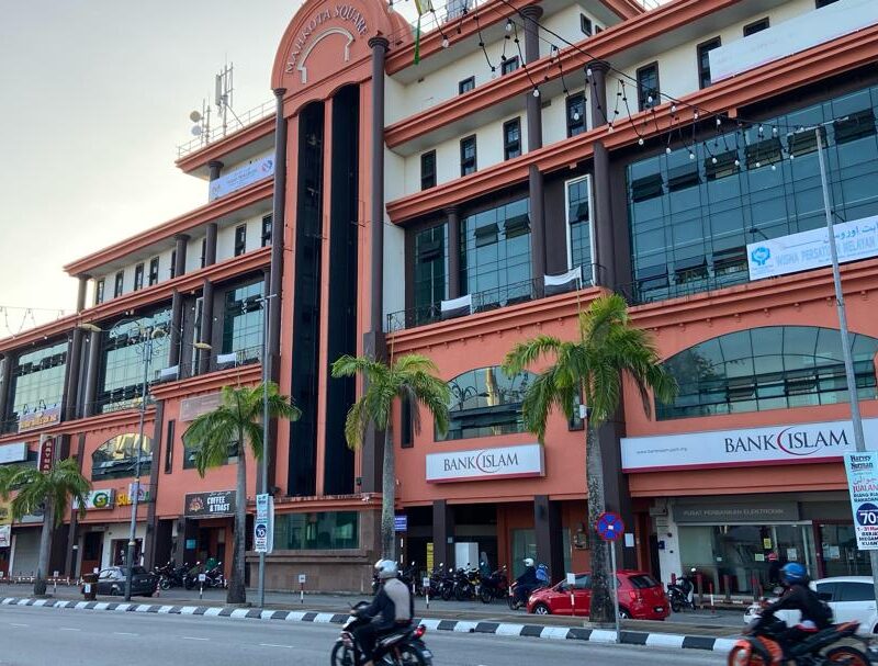 A commercial building featuring a Bank Islam storefront, palm trees, and motorcycles parked outside. The structure has a modern design with glass windows and a reddish facade, located on a busy street.