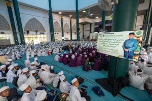 A large group of students in traditional attire sitting on a turquoise floor in a mosque hall. A banner displays information beside them, featuring a smiling man in a blue outfit. The environment is bright, with arched windows visible.