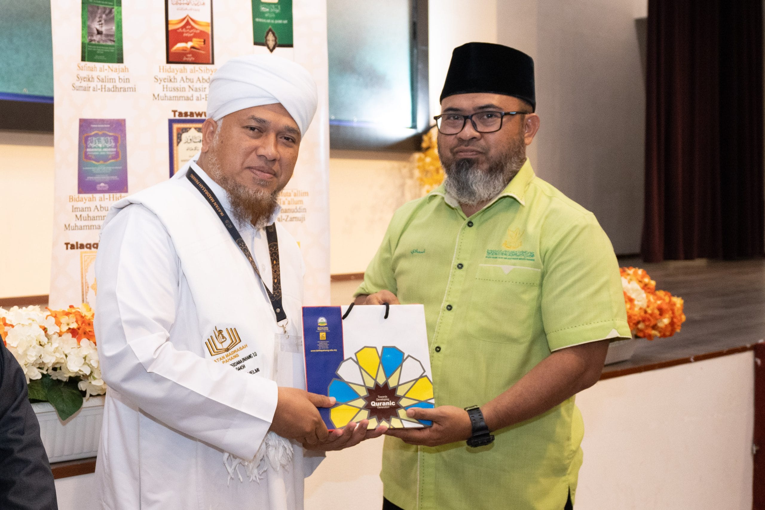 Two men standing in a hall during an event. One man in a white robe and turban presents a gift to another man wearing glasses and a green shirt. In the background, a display of books is visible.