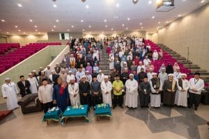 A large group of diverse participants gathered in an auditorium, posing for a group photo. They are wearing various traditional attire. The background features a section of pink seating, and tables with decorative items at the front.